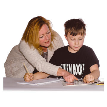 A boy sitting at a table with a book and holding a pen.  A lady is standing behind him and leaning over pointing at something in the book.