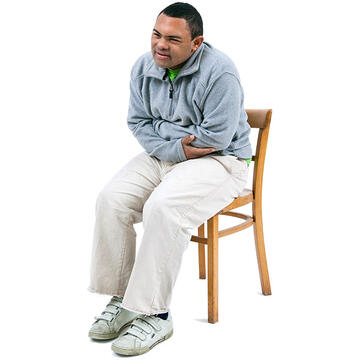 A boy sitting on a chair leaning forwards with his arms folded and looking as if he is in pain.