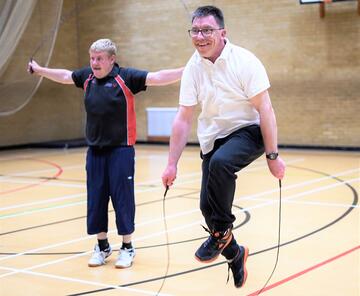 Two men in a gym. One is stretching out his arms and the other is skipping with a rope
