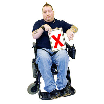 A man in a wheelchair with his thumb down and holding a piece of paper with a red cross on it.