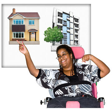 A board with a picture of  a block of flats and a house.  In front of the board is a lady in a wheelchair who is smiling and pointing to the picture of the house.e