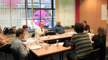 A group of people sat around a U shaped meeting table talking