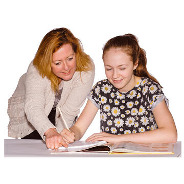 A lady standing beside a teenager who is sitting writing and looking at a book.