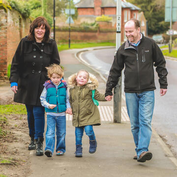A man and a woman walking along the pavement holding hands with 2 young children.