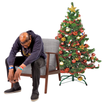 A man is slumped forward looking sad in a chair next to a Christmas tree