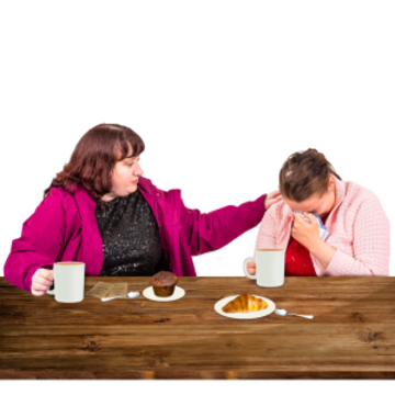 A woman is comforting a young woman who is sad whilst they share a hot drink and a cake at a table.