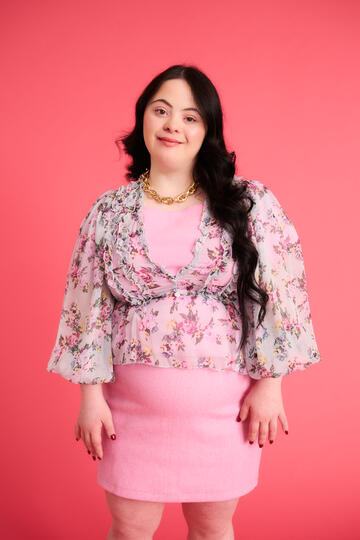 A woman wearing a light pink dress with a floral shirt over it, smiling at the camera. Behind her is a pink background