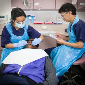 A man lying back in a dentist's chair with a dentist using instruments to look in his mouth and another member of dental staff writing.