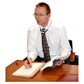 A doctor sitting at a desk holding a book and writing on an prescription pad.