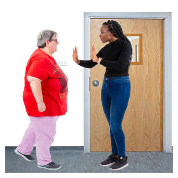 A woman walking towards a closed door, but a woman is standing in front of her with her hands out stopping her from leaving the room.
