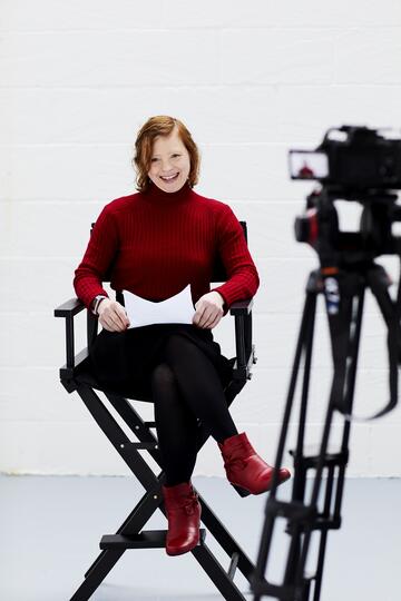 Sarah Gordy sitting on a directors chair next to a camera and reading a script