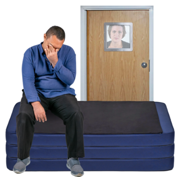 A man sitting on a mattress looking sad whilst someone watches behind a locked door