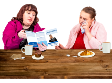 Two women sat at a table.  One woman has a tissue beside her eye. The other women is showing her a leaflet about counselling.