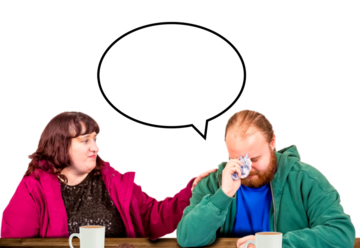 A man and a woman sitting at a table.  The woman has her hand on the man's shoulder.  The man has a speech bubble above him.  He is looking down and is wiping his eyes with a tissue.