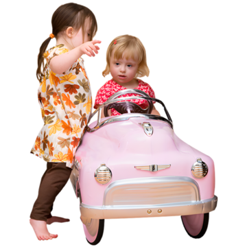 A young girl in a toy car pretending to drive it and another young girl standing beside the car and pointing.