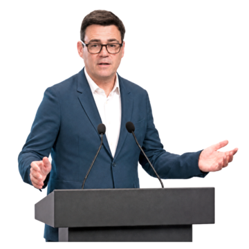 Andy Burnham wearing a blue suit and white shirt speaking at a podium.