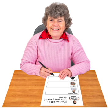Elderly woman in a pink sweater filling out an easy-read form at a wooden table.