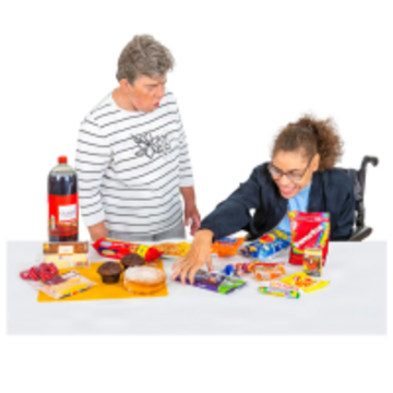 A woman reaching for a chocolate bar on a table which has cakes, fizzy drinks and other unhealthy food on it.  Another woman is looking at her.
