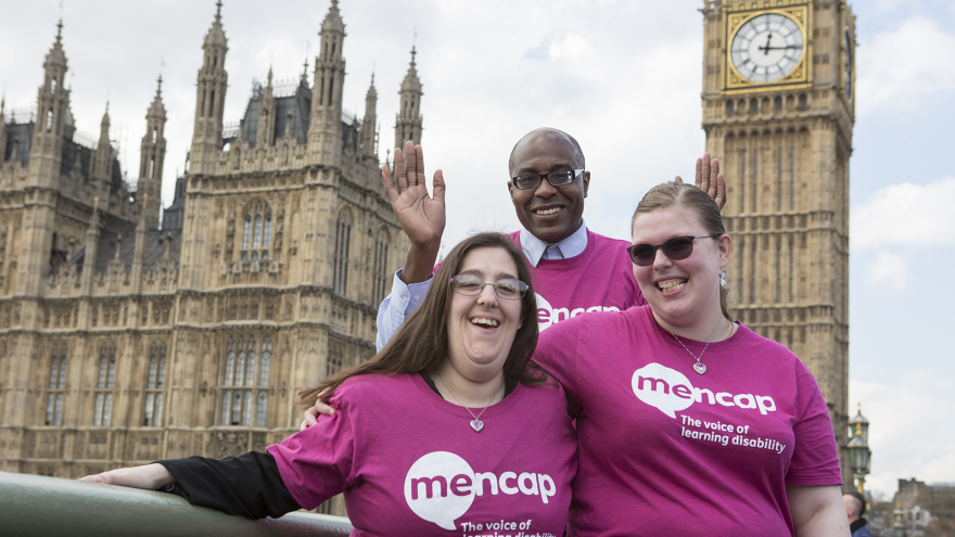 Group of three people wearing Mencap t-shirts stood in front of houses of Parliament.
