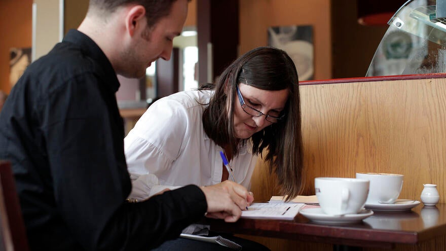 Man and woman sat at table in coffee shop. The woman is leaning over signing papers on the table.