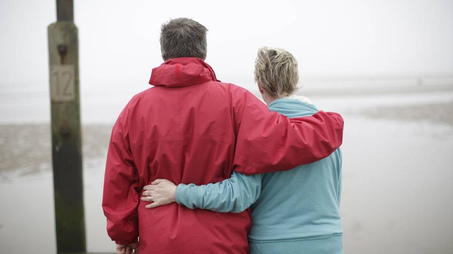 Man and woman couple with their arms around each other stood on a beach with their backs to the camera