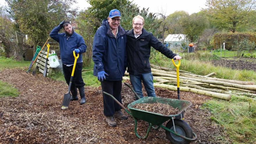 Three people stood in allotment with wheelbarrows and garden spades.
