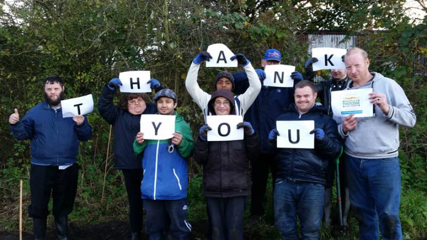 Group of people stood in garden. Each person is holding a piece of paper with a letter on it. Together the letters spell "thank you".