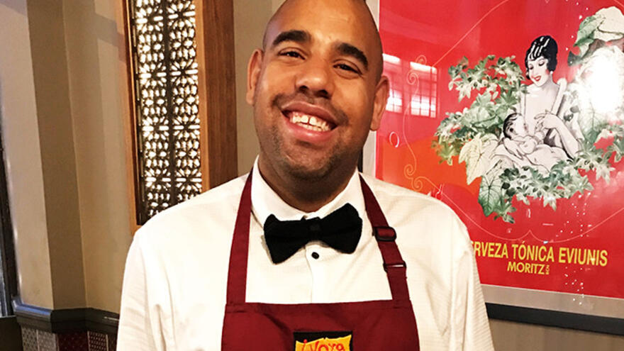 Yong man wearing shirt, bow-tie and apron smiling into camera.