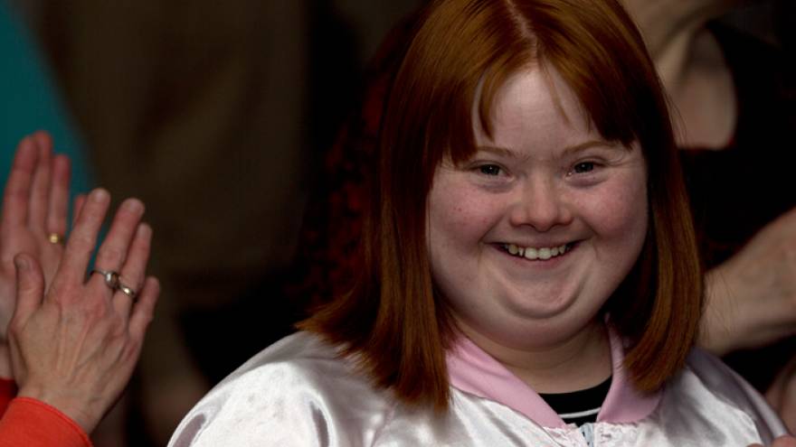 Woman with red hair wearing light pink jacket stood smiling into camera.