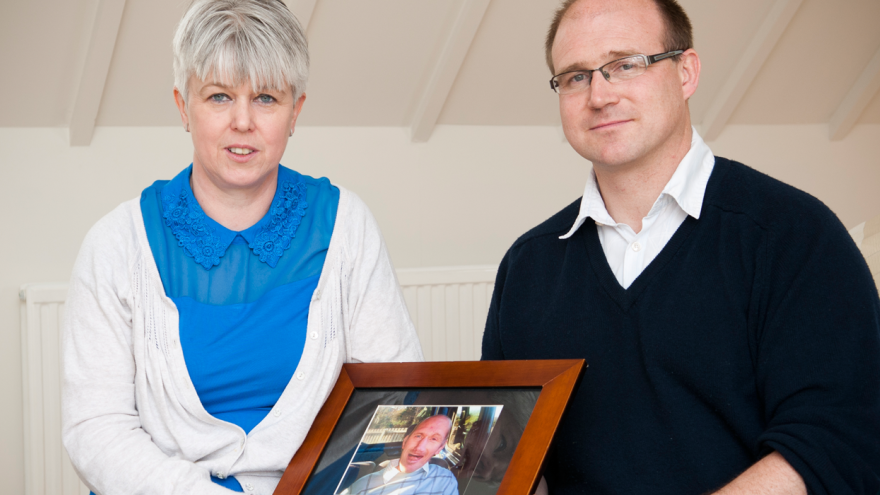 Man and woman stood in room holding framed photo of a loved one
