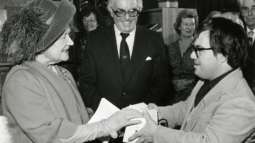 Black and white photo of Lord Rix stood behind two people who are shaking hands.