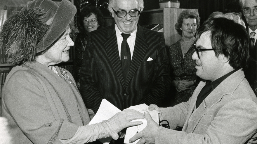 Black and white photo of Lord Rix stood behind two people who are shaking hands.