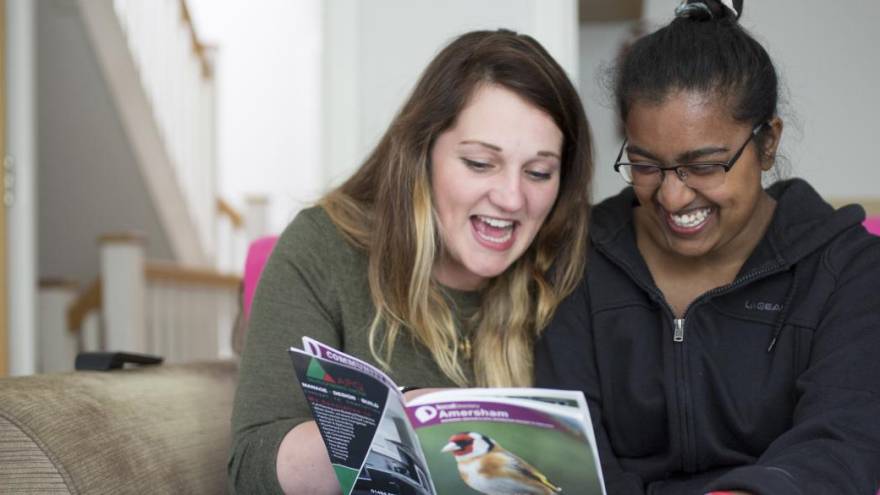 Two women sat next to each other on a sofa in a living room reading a booklet together