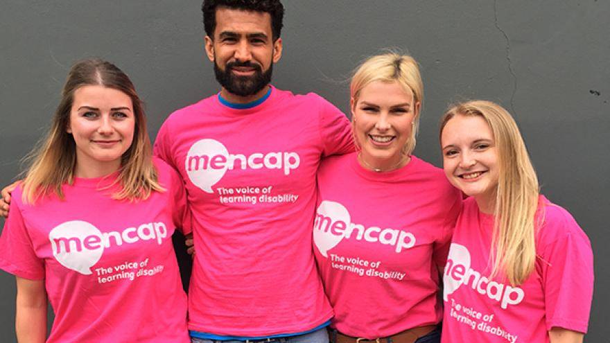 Group of four people wearing pink Mencap t-shirts stood together in front of grey wall