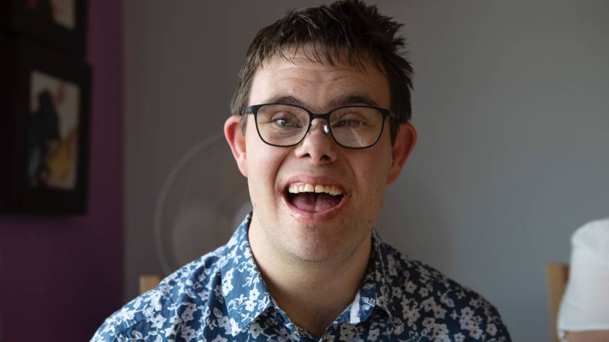 Young man with dark hair and glasses laughing into camera.