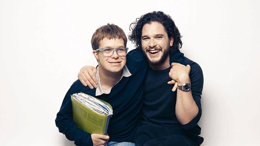 Young boy wearing glasses, holding a green book, stood next to a man with dark hair (Kit Harrington). Both men are smiling and have their arms round each other