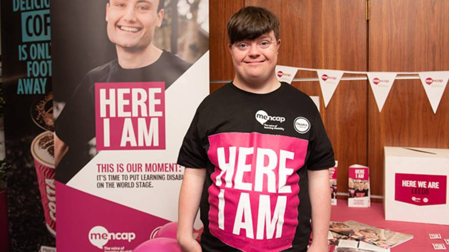 Young man, Liam Bairstow, stood in front of Mencap stand, wearing Here I Am t-shirt