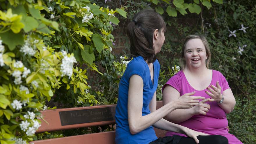 Two friends sat next to each other outside on park bench using Makaton signs to communicate. Flowering bushes in the background.