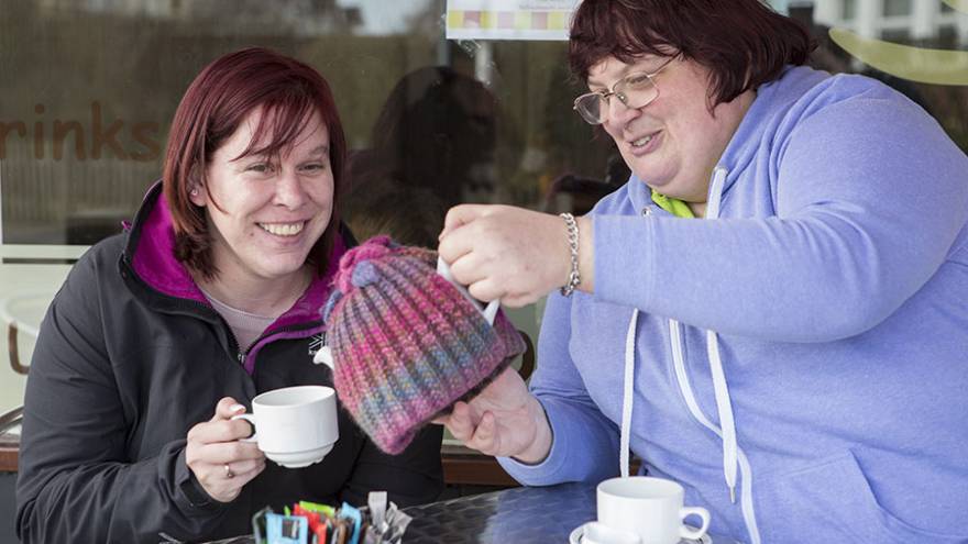 Two woman smiling sat at a table outside cafe. One woman is pouring the other tea from a teapot.