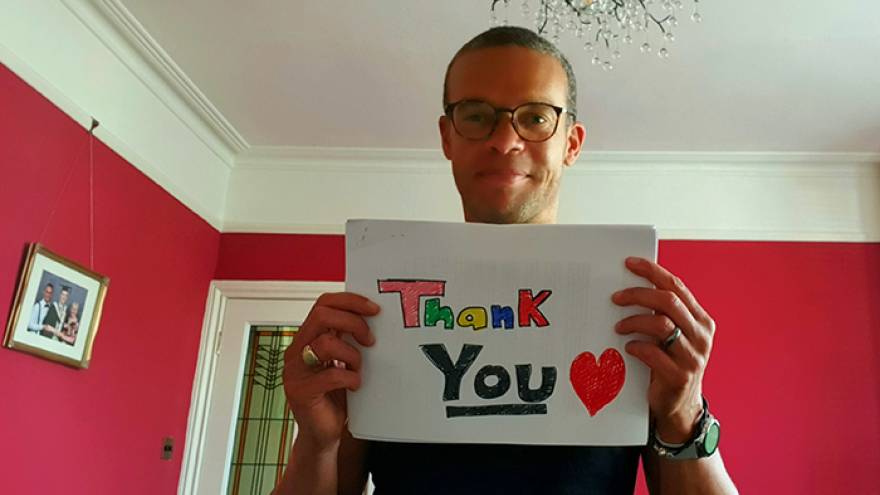 Man wearing glasses stood in living room holding hand drawn sign that reads "thank you"