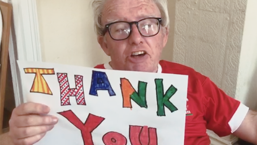 Older gentleman with glasses wearing a red t-shirt, sat holding a hand drawn sign that reads "thank you"