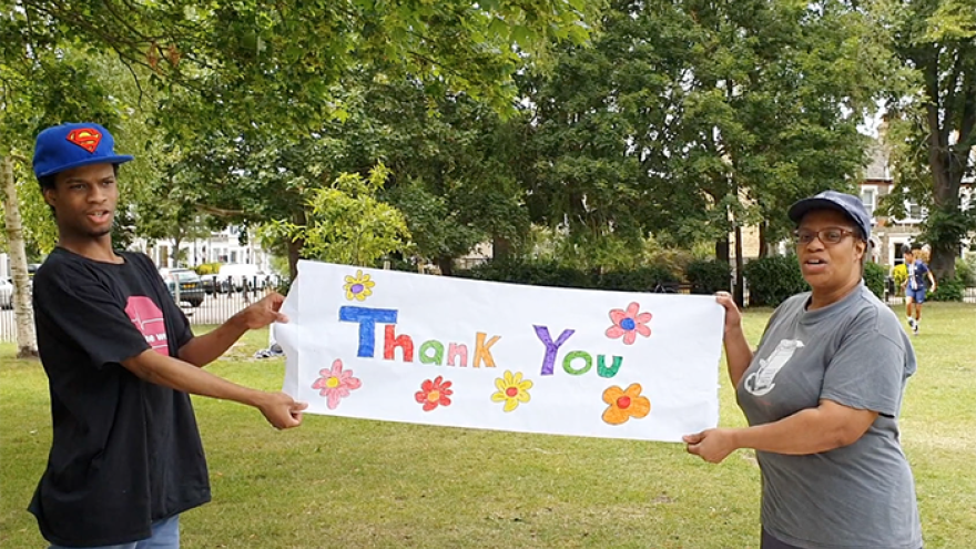 A man and a woman stood outside in a park surrounded by grass and trees, each person holding a side of a hand drawn sign that reads "thank you"