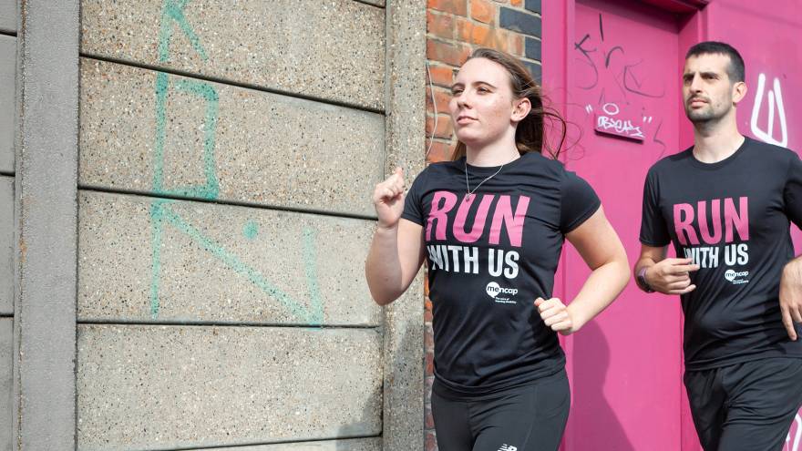 Man and woman running in front of wall whilst wearing black "Run with us" t-shirts.
