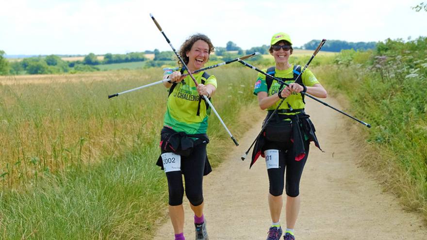Two women walking through countryside holding walking poles in front of them.