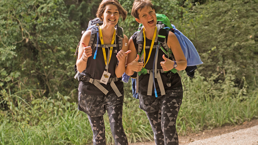 Two woman walking together through countryside, smiling and giving thumbs-up sign to the camera.