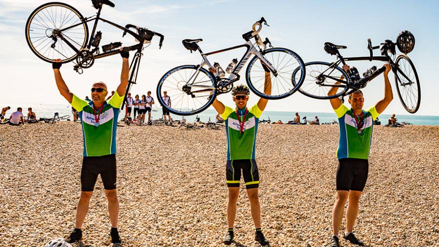 Three male cyclists stood on beach holding their bikes above their heads.