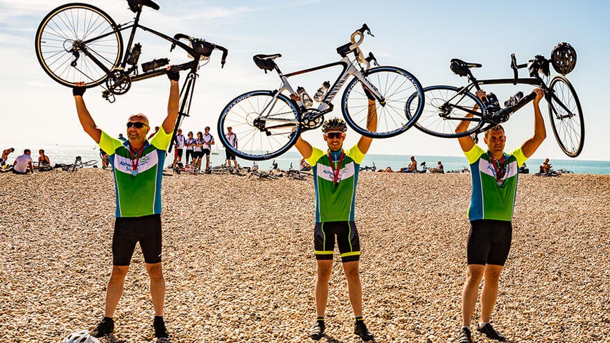 Three cyclists stood on beach holding their bikes above their heads.