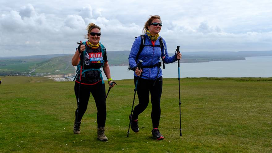 Two women walking through grass cliff-top field, sea in the background.