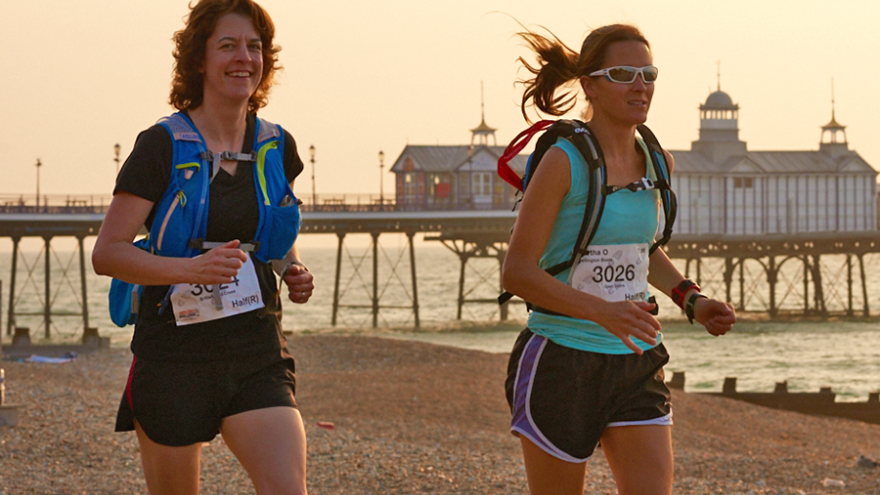 Two women running along Brighton beach at sunset. Brighton pier in the background.