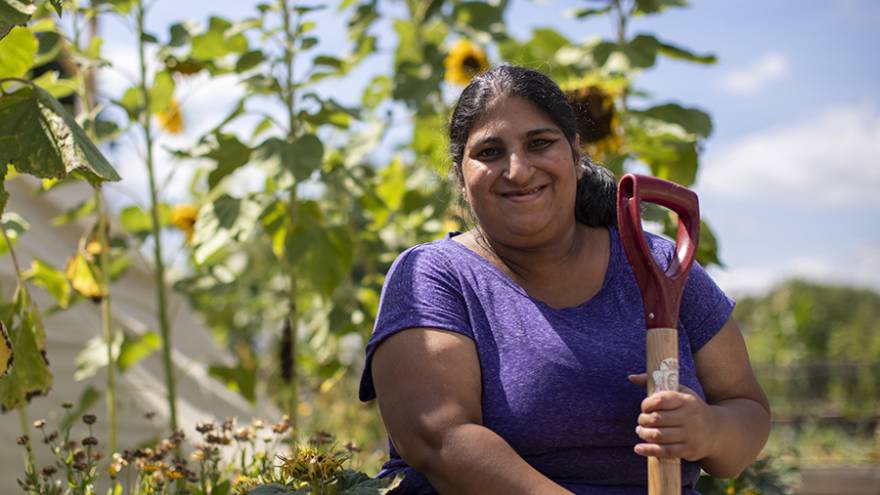Smiling woman sat outside in sunshine next to flowerbed of sunflowers, holding a spade.
